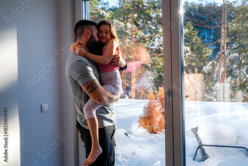 Father is holding his young daughter in his arms, embracing her affectionately by a window overlooking a snowy winter landscape, creating a heartwarming scene of familial love and warmth