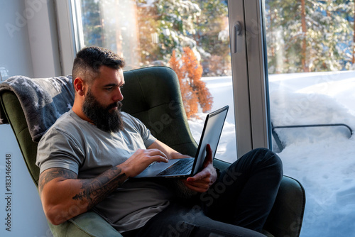Freelancer with tattoos and a beard working on a laptop in an armchair by a window, enjoying the warmth and snowy landscape during winter
