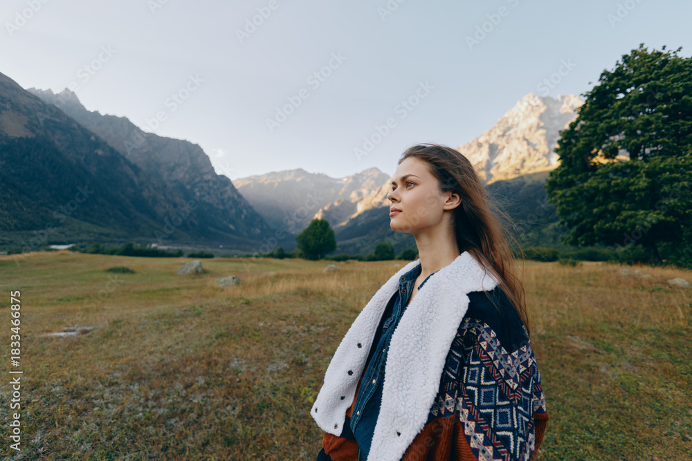 Fototapeta premium Woman portrait in mountains meadow, nature outdoors and travel landscape setting with thoughtful expression, cozy patterned coat and wide valley view at golden hour.