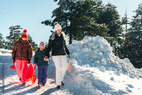 Mother and her two daughters are enjoying a winter day, walking on a snowy mountain path with their sleds, surrounded by snow-covered trees and a bright blue sky