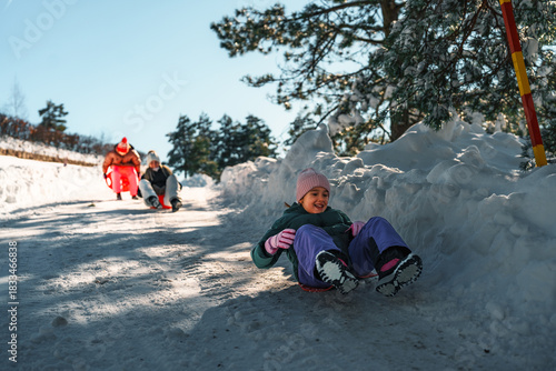 Smiling girl enjoying winter sledding fun, sliding downhill on a snowy slope, with her family following behind on a bright sunny day, creating a joyful winter scene