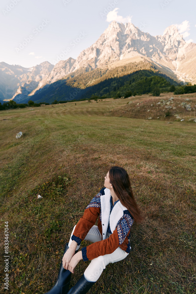Fototapeta premium Woman in autumn sweater sits in a mountain meadow, nature landscape with high peaks and soft light, travel and hiking scene, peaceful outdoor solitude and scenic horizon.