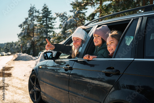 Mother and daughters enjoying winter road trip pointing at something in the distance while traveling by car on a snowy road surrounded by snow-covered trees