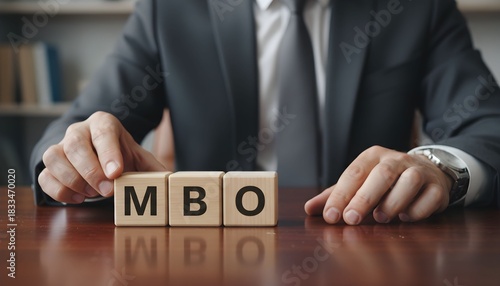 Businessman in a suit at a wooden desk arranging blocks to spell out MBO for management buyout.