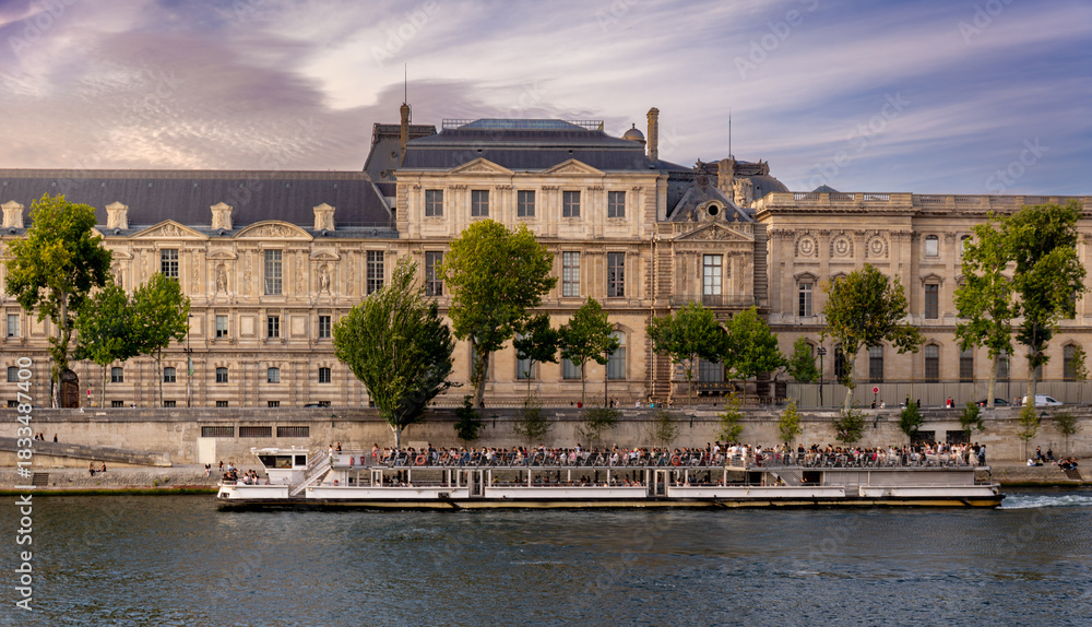 Fototapeta premium A tourist boat on waters of Seine River in Paris, France, in front of the Louvre Museum building with tourists enjoying the cruise along Paris's most beautiful historical axis