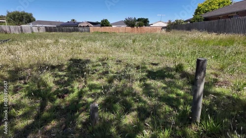 A vacant residential lot in an Australian suburban neighbourhood, featuring tall grasses and open land bordered by timber posts and fences. Concept of land availability, and unmaintained green space
