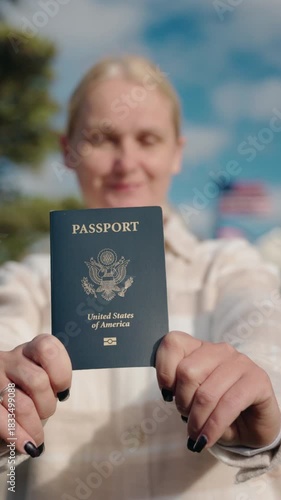 Woman holding a United States passport outdoors with an American flag waving in the background on a bright sunny day
