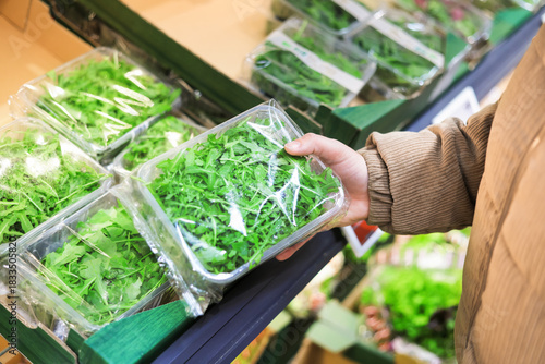 Canvas-taulu Customer holding a plastic container of fresh green arugula leaves while shopping in the supermarket produce section