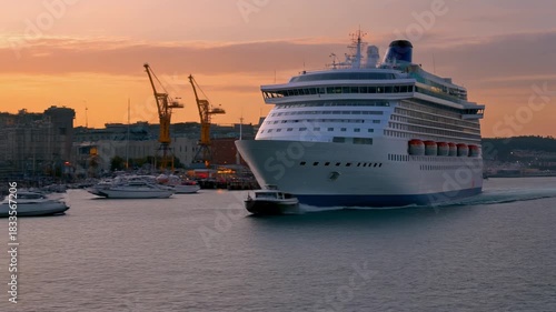 A large white cruise ship sailing in a harbor with smaller boats and port cranes in the background during sunset.