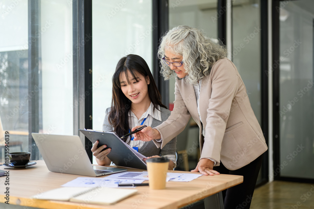 Fototapeta premium Old asian businesswoman looking at clipboard talking helping young coworker at table in office room
