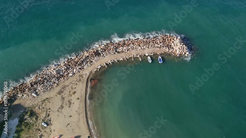 Aerial drone view of a curved rocky breakwater surrounded by clear turquoise sea water. Small boats anchored near the coast with gentle waves creating natural patterns