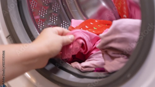 Close-up of a person hands putting or taking out pink and red laundry into the open door of a front-loading washing machine.
