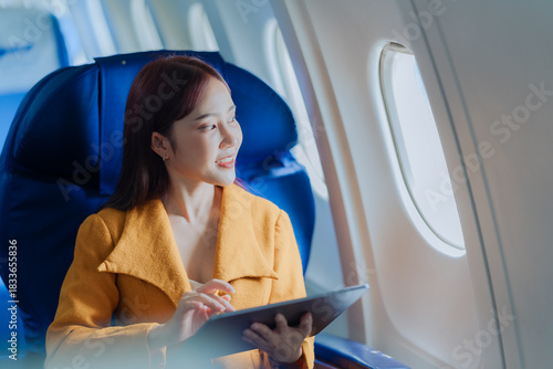 Asian woman using digital tablet while traveling by plane. Female passenger using digital device in cabin near window. She is traveling for business.