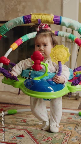 Baby girl standing and bouncing in a colorful activity jumper, holding bright toys and exploring them with curiosity