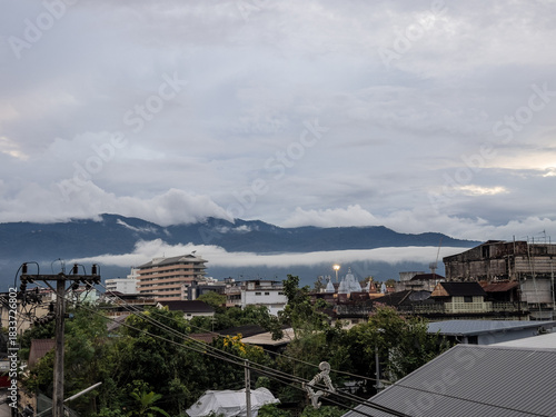 Overcast morning in Chiang Mai, Thailand, with a city skyline featuring diverse architecture, power lines, and lush mountains shrouded in mist and clouds. Urban sprawl meets natural grandeur.