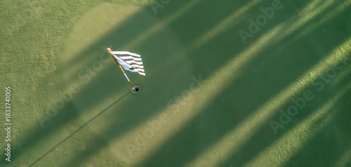 Aerial view of golf flag on green course with long shadows cast by trees. vibrant green grass contrasts beautifully with black and white flag, creating serene golfing atmosphere