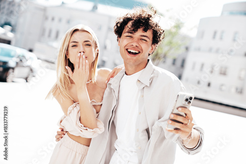Young smiling beautiful woman and her handsome boyfriend in casual summer clothes. Happy cheerful family. Female having fun. Couple posing in street. Hold smartphone, use phone apps, look at screen