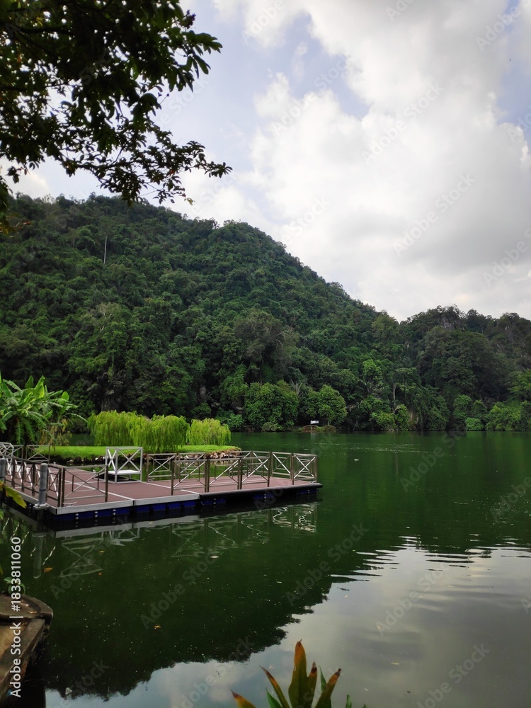 Fototapeta premium A wooden floating dock extending into a calm green lake backed by a dense, forested hill. The scene is tranquil and quiet. Use it for lifestyle websites, travel campaigns, or environmental reports.