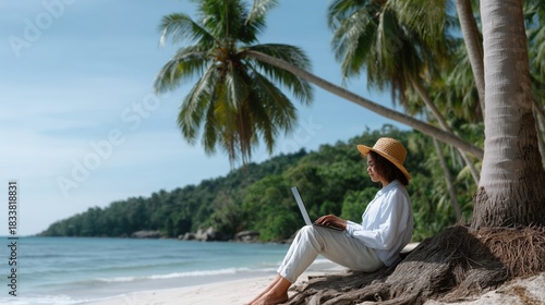 Fototapeta Naklejka Na Ścianę i Meble -  Woman is sitting on the beach with a laptop. She is wearing a straw hat. The beach is near a palm tree