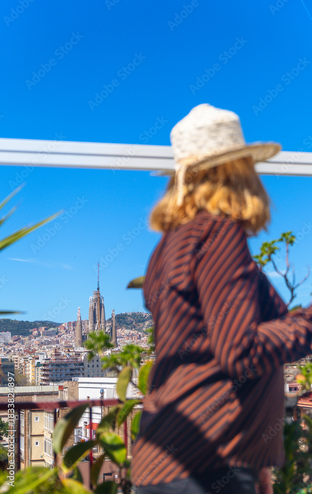 Obraz premium Woman with her back to the camera, wearing a straw hat, on a sunny rooftop overlooking the city of Barcelona, with the Sagrada Familia in the background.