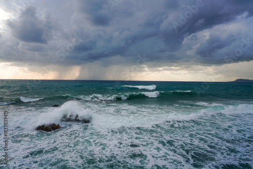 Dramatic view of strong wind and high waves crashing over rocks under a dark, brooding storm sky. Intense marine landscape with a visible rain squall on the horizon.