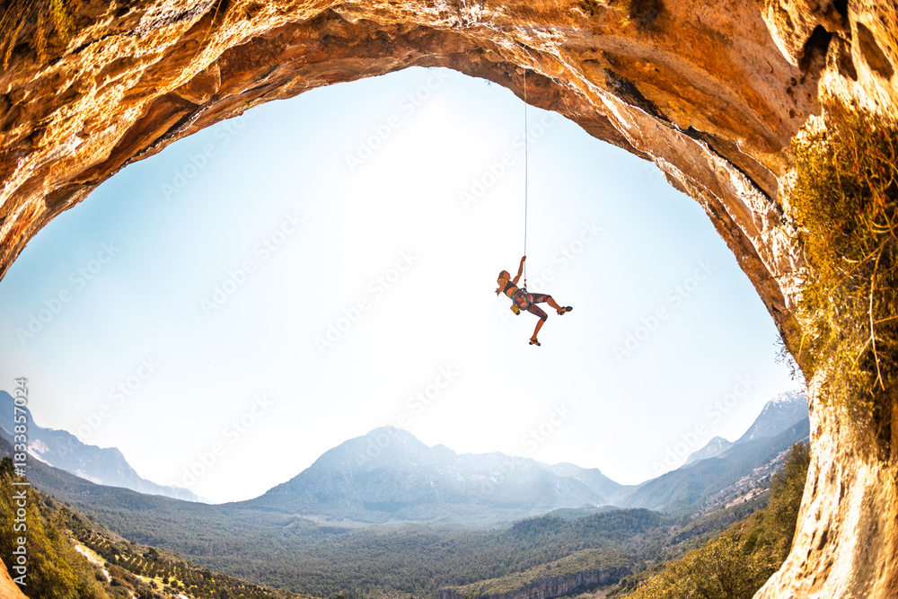 Fototapeta premium A climber hangs on a rope. A female climber descends. Rock climbing in Turkey. A woman engaged in extreme sports. An arched rock face. A climbing route in a cave.