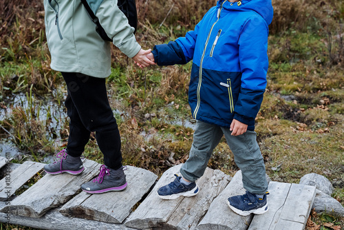 siblings exploring nature together, two young explorers with colorful clothing on woodland pathway, brothers and sisters carefully traverse mossy boardwalk amid puddles and fall scenery