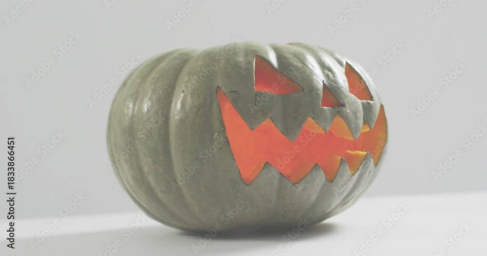 Fototapeta premium Glowing carved green-gray pumpkin casting warm orange light on white tabletop, pale gray wall