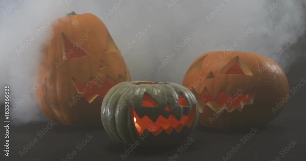 Fototapeta premium Glowing green-gray jack-o'-lantern sitting on studio table lit, fog drifting around pumpkins