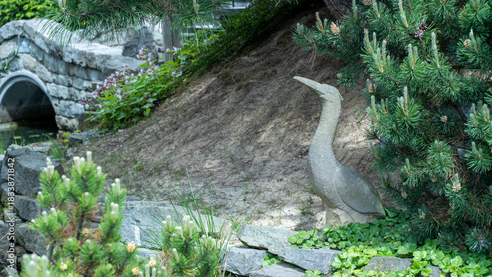 Αυτοκόλλητο Stone bird statue in lush garden with conifer trees and rock bridge