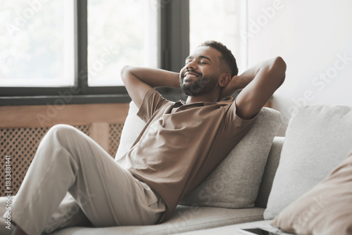 Satisfied handsome young man relaxing on sofa at home in living room, resting after a hard day work, closed eyes, put hands behind head, smiles happily. Relaxation, holiday, enjoy life concept