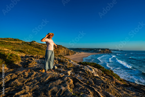 Beautiful middle-aged woman tourist standing on cliff overlooking Monte Clerigo beach and ocean in summer. Algarve coast in Portugal. Side view