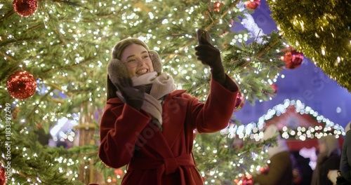 A young beautiful girl in a red coat walks in the evening at the European Christmas Market making a video call standing against the backdrop of a Christmas tree with garlands and illuminations