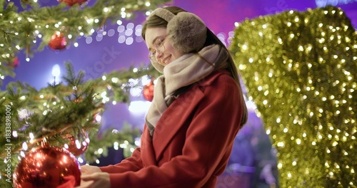 A young beautiful girl in a red coat stands near a Christmas tree with garlands and illuminations in the evening at the European Christmas Market and touches the Christmas tree decorations.