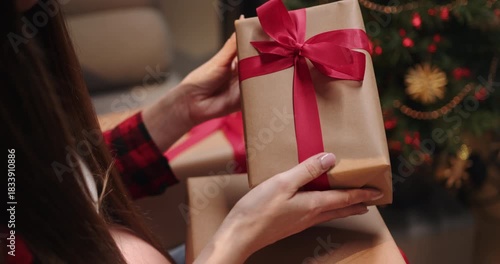 A person is holding a wrapped present with a red ribbon in front of a Christmas tree. A person is holding wrapped presents near a decorated Christmas tree, creating a festive holiday scene
