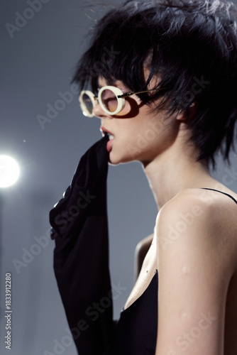 Stylish woman in black outfit biting sleeve wearing round glasses with tousled short hair against gray background.