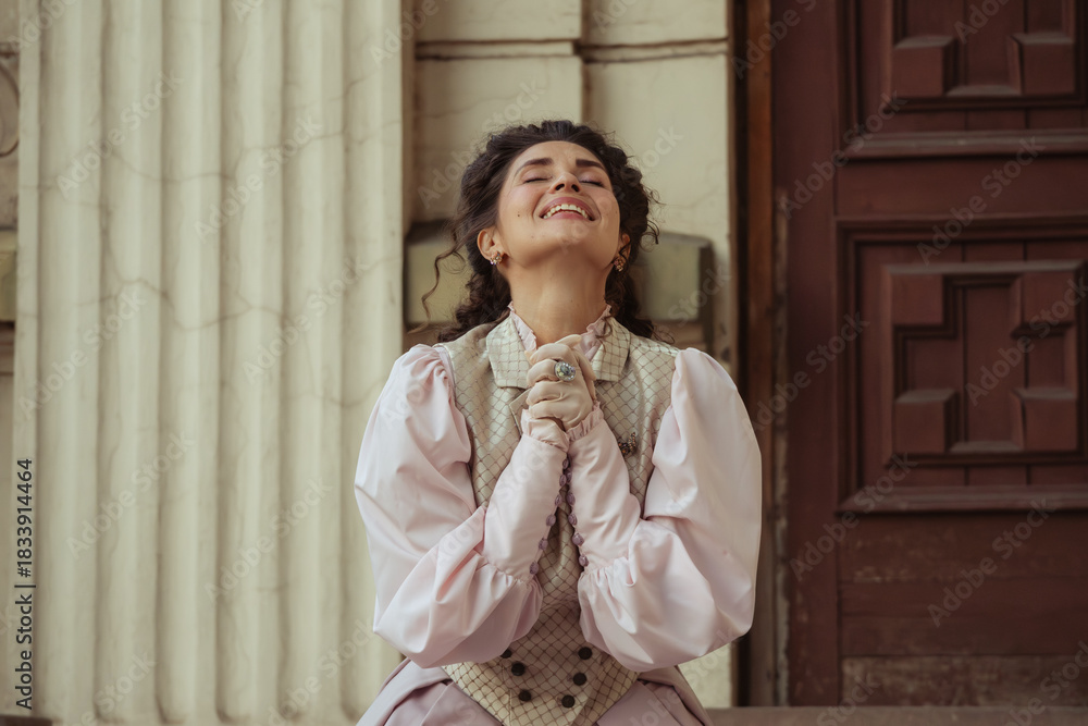 Fototapeta premium An elegant woman in a vintage dress sits next to an old stone column with a very happy expression on her face.