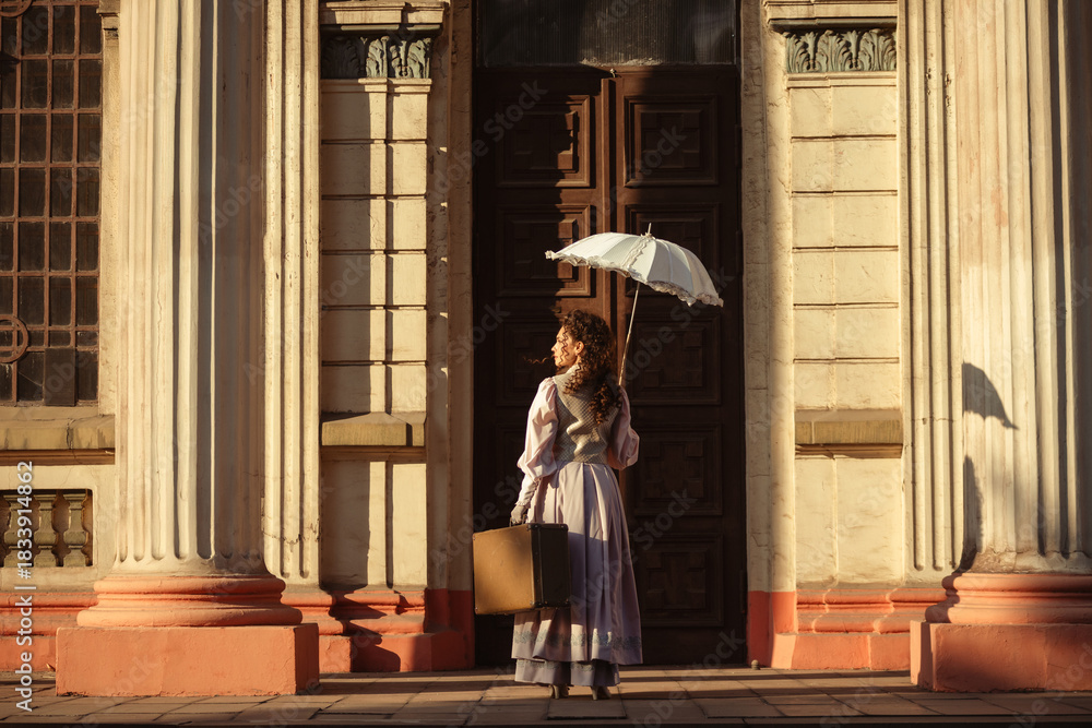 Obraz premium A woman in an antique dress with an umbrella and a suitcase stands in front of the doors of an old cathedral.