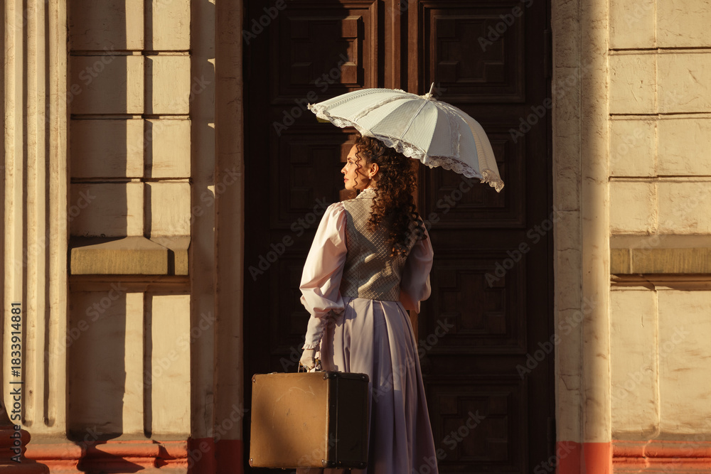 Fototapeta premium A woman in an antique dress with an umbrella and a suitcase stands in front of the doors of an old cathedral.