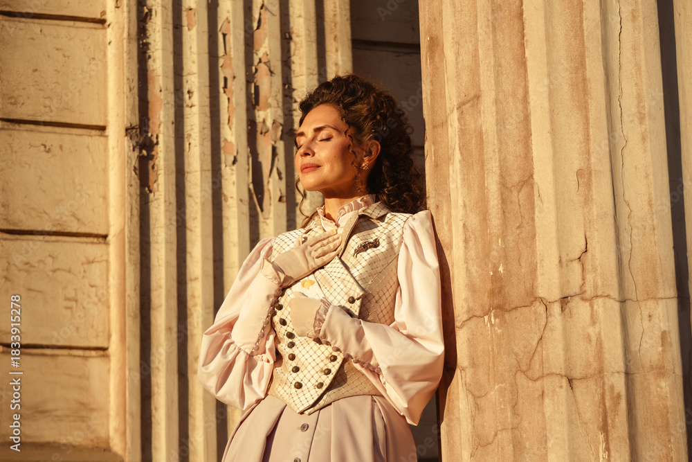 Fototapeta premium An elegant woman in a vintage dress stands by an old stone column