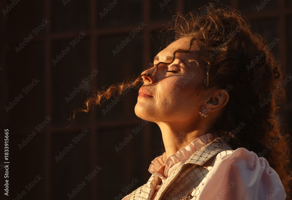Obraz premium Portrait of a woman in vintage clothing, illuminated by warm sunlight, against a backdrop of ancient architecture.