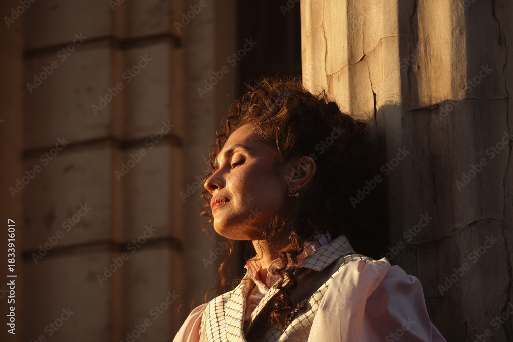 Obraz premium Portrait of a woman in vintage clothing, illuminated by warm sunlight, against a backdrop of ancient architecture.