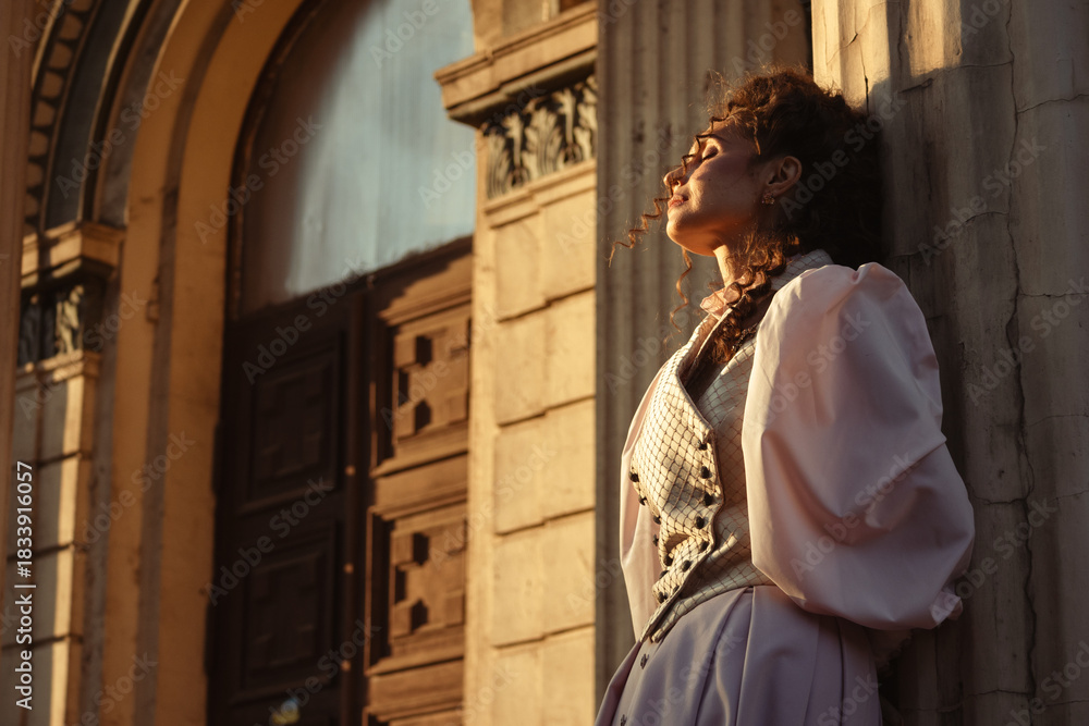 Obraz premium Portrait of a woman in vintage clothing, illuminated by warm sunlight, against a backdrop of ancient architecture.