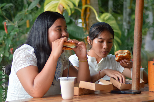 A close-up of two teenage girls sitting at a wooden table in an outdoor garden cafe, enjoying a meal. One takes a large bite of her burger while her friend adjusts hers, with a drink nearby.
