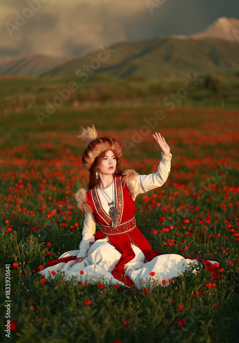 Young woman in Kyrgyz traditional clothing red and white sitting in field of poppies on mountain background. Female adult in national dress