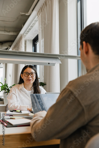 A female worker laughs and talks to a male worker across from her while they sit at a table in front of laptops