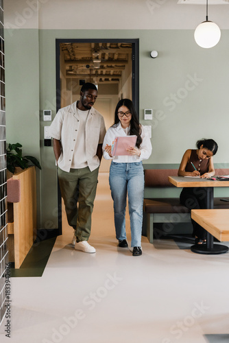 A male and female workers are reading a notebook she is holding as they walk past a female worker who is sitting at a desk and writing in a notebook