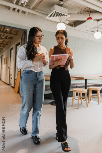 Two female workers walk and read a notebook that one of them is showing and talking