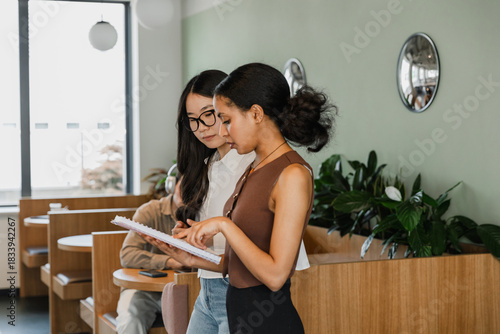 A female worker is talking and pointing at a notebook which she is showing to a female worker walking next to her
