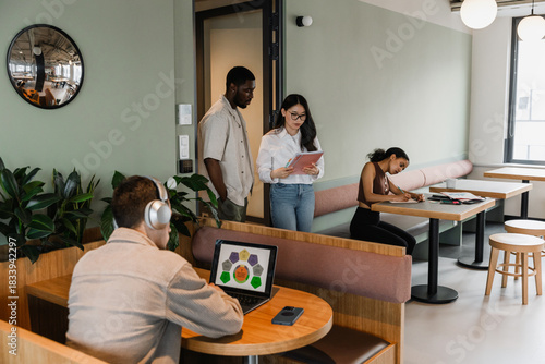 Male and female workers reading a notebook and walking past two workers sitting at desks while writing in a notebook and looking at a laptop